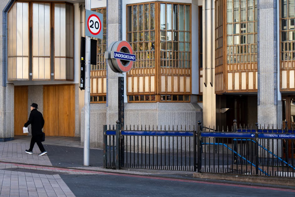 An exterior view of a modern building with large wooden lattice window panels and grey concrete walls, adjacent to an underground station entrance marked by a round 'Underground' sign and a blue raili